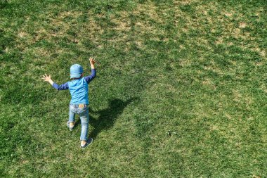 Mavi şapkalı küçük çocuk, güneşli bir günde bahar parkında yemyeşil çimlerle kaplı dik yamaçtan yukarı çıkar.