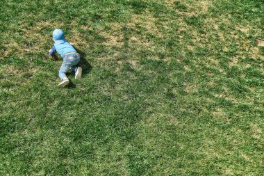 Mavi şapkalı küçük çocuk, güneşli bir günde bahar parkında yemyeşil çimlerle kaplı dik yamaçtan yukarı çıkar.
