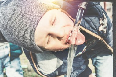 Gri örgü şapkalı komik küçük çocuk güneşli bir günde köy evinin bahçesindeki su pompasından su içer.