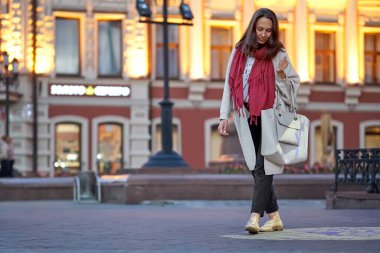 Attractive young long haired woman wearing stylish light coat and bright pink scarf walks along old city street at sunset
