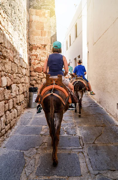 Tourists riding donkeys equipped with leather saddles on weathered narrow paved road on Lindos historical town street in Greece