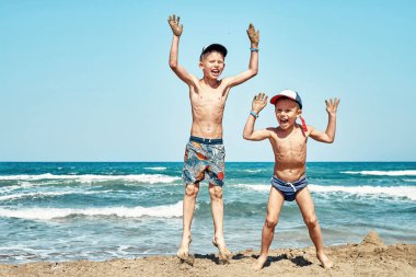 Şortlu neşeli küçük kardeşler kumsalda ıslak kumsala atlarlar. Gök mavisi dalgalara ve yaz günü Prasonisi pelerinine karşı.