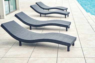 Geri dönüştürülmüş malzemeden yapılmış yeni siyah hasır plastik şezlonglar parlak güneş ışığı altında, havuz kenarında, aydınlık fayanslı zemin üzerinde duruyor.