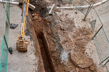 Kovalı renkli kazıcı, çitlerle çevrili yeni su borularını değiştirmek için derin çukurun yanında duruyor.
