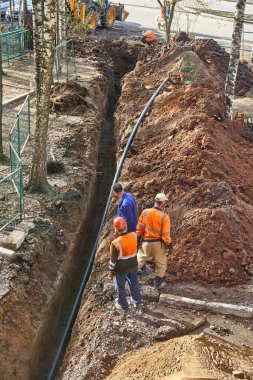 İzhevsk, Rusya - 08 Mayıs 2021: Üniformalı işçiler bahar günü konut sahasında yeni plastik su borusuyla dar bir siperin yanında duruyorlar