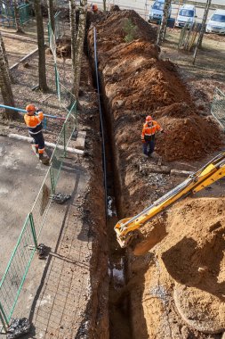 İzhevsk, Rusya - 08 Mayıs 2021: Çağdaş sarı kazıcı güneşli bahar gününde konut inşaatı alanındaki su boru hattının değiştirilmesi sırasında dar çukurlar kazıyor