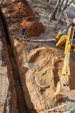 İzhevsk, Rusya - 08 Mayıs 2021: Çağdaş sarı kazıcı güneşli bahar gününde konut inşaatı alanındaki su boru hattının değiştirilmesi sırasında dar çukurlar kazıyor