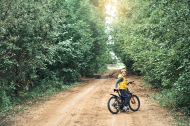 Sarı yelekli, modern bisikletli sevimli küçük çocuk yaz aylarında yeşil ağaçlı kırsal yoldaki kameraya bakıyor.