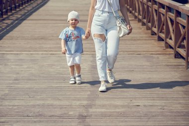 Beyaz şapkalı sevimli çocuk, güneşli yaz gününde genç annesiyle el ele tutuşarak ahşap yaya köprüsünde yürüyor.
