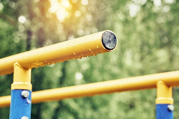 春のスポーツグラウンドで雨滴をぶら下げ練習のための黄色の金属バー曇りの日に緑の公園極端な近景 — ストック写真