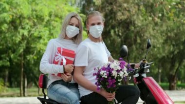 Bir adam ve koruyucu maskeli bir kız bir motosiklete binip kameraya bakıyorlar. Kız beyaz kurdeleli kırmızı bir kutuya sarılıyor ve adam elinde bir buket çiçek tutuyor..