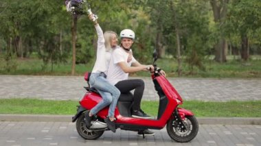 Elinde çiçek buketi olan bir kız ellerini kaldırıp kırmızı bir motosikletin üzerinde bir adamın yanında oturmaktan çok mutlu olur..