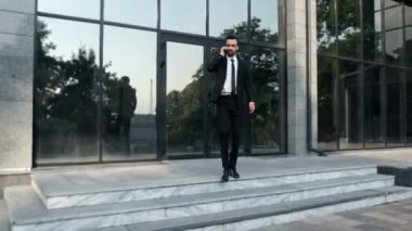 Takım elbiseli bir adam, telefonla konuşuyor, camdan pencereleri olan bir binadan çıkıp, çevreyle dost olan bireysel aracına doğru yürüyor..