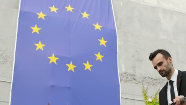 ビジネス服の男は 欧州連合の旗の背景にスクーターでフレームに入り 旗を見て 楽観的に笑顔 — ストック動画