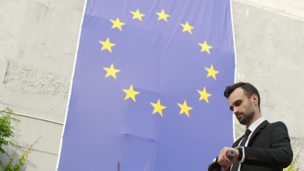 一名身穿西装的男子在欧洲联盟旗帜的背景下骑着一辆踢踏车停下 看着国旗 转身看着摄像机 — 图库视频影像