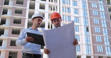 Tabletli bir mühendis, inşaatçıya kağıt ve elektronik çizimleri gösterir ve inşaat halindeki bir konut binasına işaret eder..