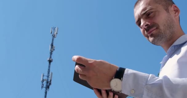 一个男人站在通信塔的后面 把他的平板电脑和5G网络连接起来 — 图库视频影像