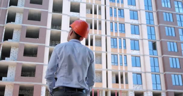 オレンジの安全ヘルメットの建設労働者は 建設中の住宅の建物を見て 紙の図面を展開し それらを見て その後 建設現場で — ストック動画