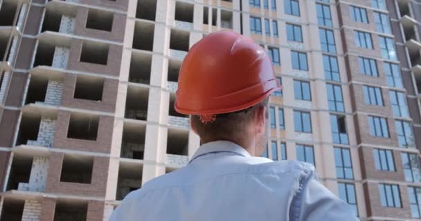 オレンジ保護ヘルメットの労働者がその場所から建設中のアパートを検査する — ストック動画