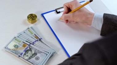 Bir adam kalemle Bitcoin 'in değerini yazar, ki bu da söz konusu, panodaki bir kağıt parçasına. Masanın yanında para ve bitcoin var..