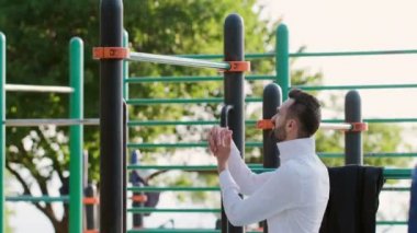 Beyaz tişörtlü bir adam kollarını uzatıyor, kendini bara çekiyor, sokak parkında ofis kıyafetleriyle spor yapıyor..