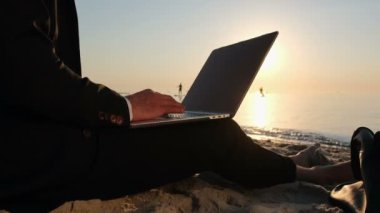 Bir iş adamı, sabahın erken saatlerinde deniz kenarındaki kumsalda takım elbisesiyle oturup dizüstü bilgisayarda çalışır..