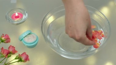 Elleri ile bir gül çiçeğinin yapraklarını cam bir kapta suya indirir, kasenin yanında bir buket gül, küçük bir kavanoz el kremi ve bir mum yanar..