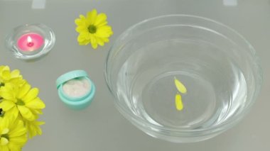 Sarı çiçek yaprakları suyla dolu cam bir kaseye düşer. Masanın üzerinde sarı çiçekler, açık bir el kremi kavanozu ve yanan pembe bir mum var..