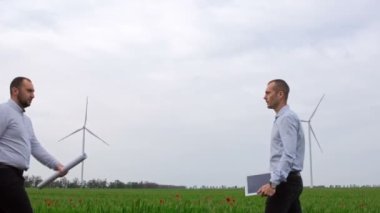 Elinde tablet ve kağıt olan iki adam yeşil bir alanda çalışan rüzgar çiftliklerinin arka planına karşı el sıkışıyorlar. Yeşil enerjiye yatırımlar.