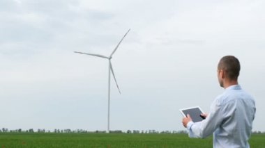 Tabletli bir yatırımcı yel değirmeninin arka planında dikiliyor ve kameraya doğru dönüyor..
