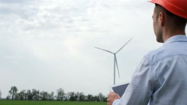 Elinde tabletle koruyucu kasklı bir adam çalışan rüzgar tarlalarını inceliyor. Rüzgardan çevre dostu elektrik üretimi.