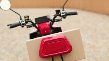 Kırmızı tişörtlü bir teslimatçı, kırmızı elektrikli motosikletli bir karton kutuda bir paket alıyor. Elektrikli taşımacılık ile küçük paketler teslim.