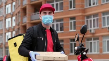 Kırmızı şapkalı ve virüs korumalı bir adam iki kare karton kutuyu kameraya doğru tutuyor ve başparmağını kaldırıyor..