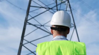 Üniformalı bir güç mühendisi yüksek voltajlı bir güç hattına doğru yürür, kamera mühendisi arkadan yakalar..