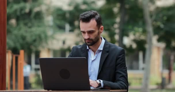 スーツ姿の男がノートパソコンで働き コーヒーを飲みながら路上のカフェに腰を下ろしている インターネットワーカーのためのコーヒー — ストック動画