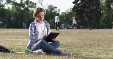 Sarışın kadın yeşil çimenlikte modern tabletle ödev yapıyor.