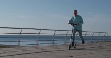 Kot pantolonlu ve kapüşonlu bir adam kaldırımda elektrikli scooter kullanıyor.