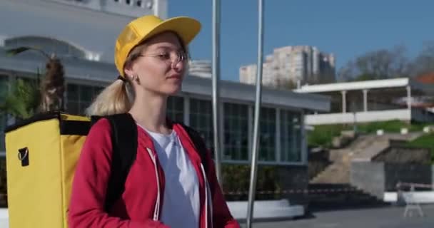 Блондинка-курьер с фирменным рюкзаком и желтой кепкой — стоковое видео