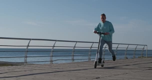 Uomo in jeans e felpa con cappuccio cavalca scooter elettrico sul lungomare — Video Stock