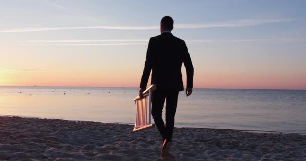 L'homme marche le long de la plage de sable et s'assoit sur une chaise pliée en bois — Video