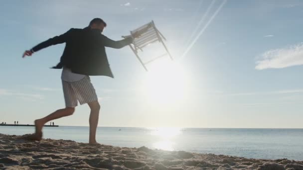 Arg kille i kostym och shorts sätter stol på sand och sitter — Stockvideo