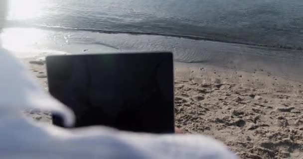 一个男人坐在海边靠近水边的笔记本电脑上工作 一只狗沿着海滨跑着 拍拍着一个男人的肩膀 — 图库视频影像