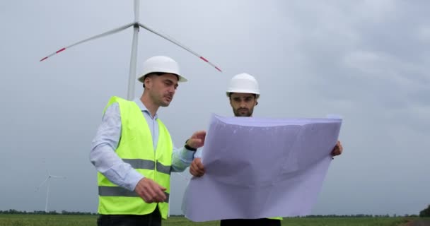 身穿防护服的工程师站在多云天气下工作的风车的背景下 讨论风车的工作 一位工程师正在拿着一张大纸图纸 — 图库视频影像