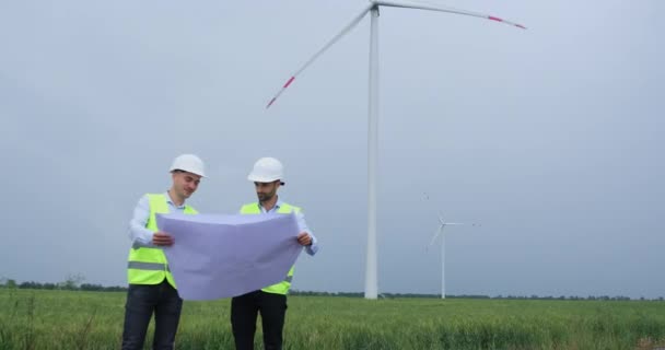 两名身穿制服的工程师在风力涡轮机可再生能源的背景下讨论使用一个大蓝图的问题 — 图库视频影像