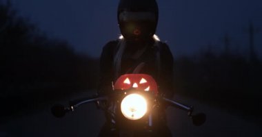 Bir bayan bisikletçi, gece farları açık bir şekilde motosikletin üzerinde otururken önünde bir Cadılar Bayramı balkabağı feneri tutuyor. Azizlerin günü yaklaşıyor..
