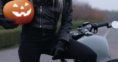 Kasklı bir kadın bisikletçi elinde parlayan gözleriyle cadılar bayramı balkabağı tutarken yolda motosikletinin yanında duruyor..