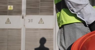 Elektrik mühendisi, bir elektrik santralinin önünde dikilirken arkasında turuncu bir koruyucu kask tutuyor..