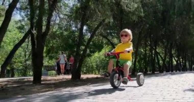 Güneş gözlüklü küçük çocuk kaldırımlı yolda üç tekerlekli bisiklet sürüyor.