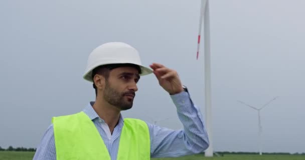 Инженер надевает белый шлем и смотрит на ветряную турбину — стоковое видео