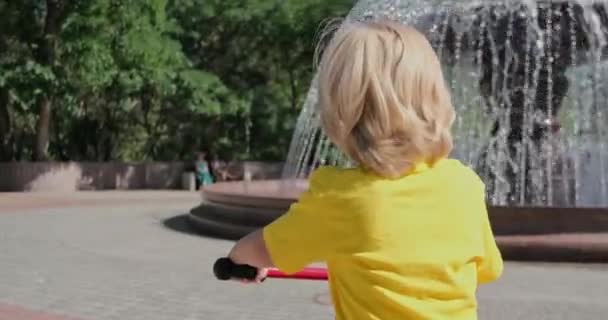 Bambino con capelli biondi cavalca triciclo guardando fontana — Video Stock